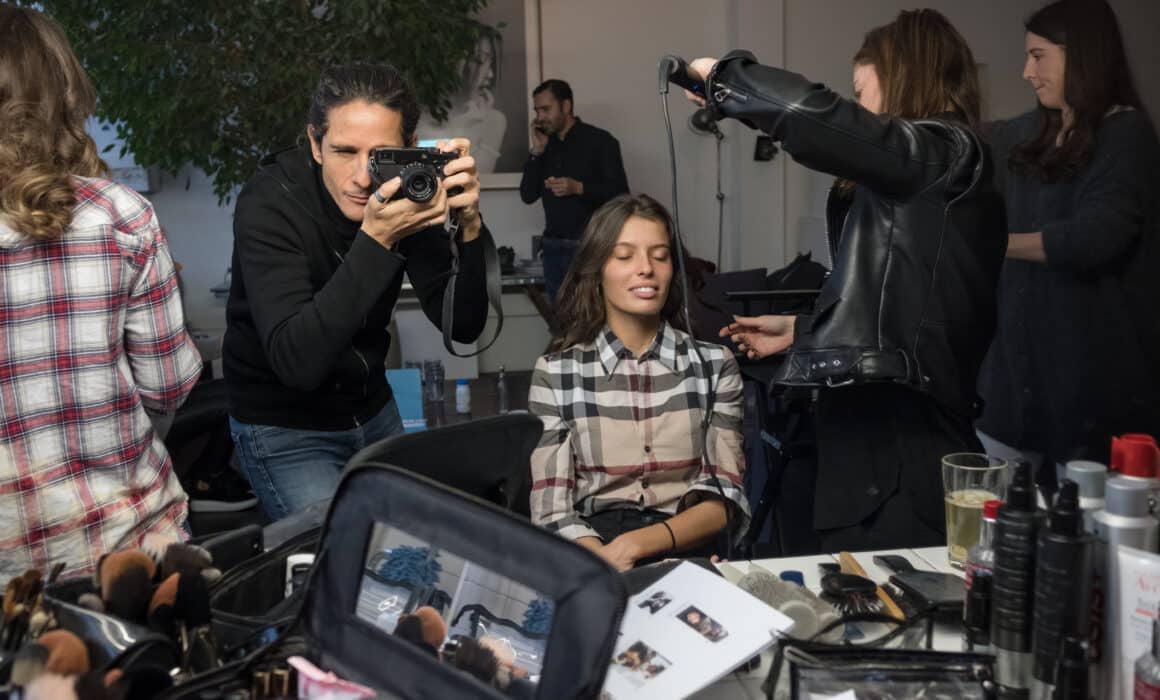 Vue des coulisses d'un shooting de mode montrant un photographe prenant une photo d'un mannequin assis, pendant qu'une coiffeuse s'occupe de ses cheveux. Au premier plan, une table remplie de produits cosmétiques et de pinceaux de maquillage.