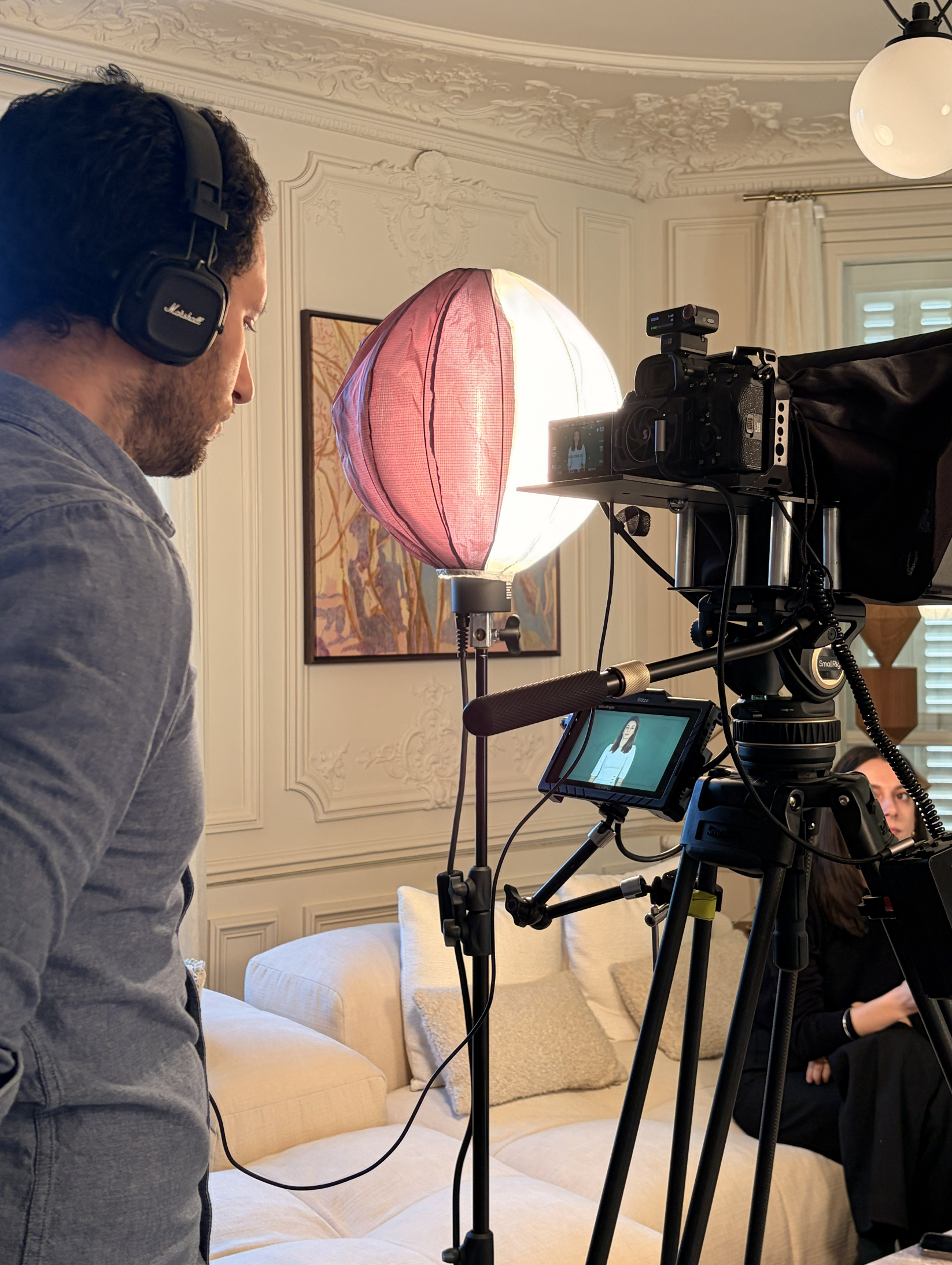 tournage-video-corporate-photographe-professionnel Coulisses d'un tournage vidéo corporate dans un salon avec un vidéaste professionnel équipé d'une caméra sur trépied, d'un moniteur de contrôle et d'un éclairage de studio.